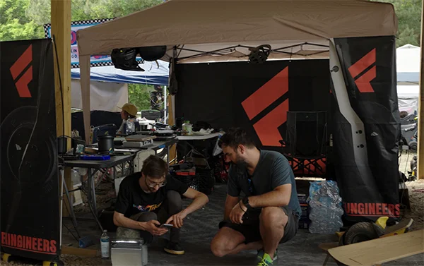 The Fungineers booth at Oak City Shred Fest, a canopy tent hung with black banners carrying the red Fungineers logo, with two people crouched over a phone beside boards and gear.