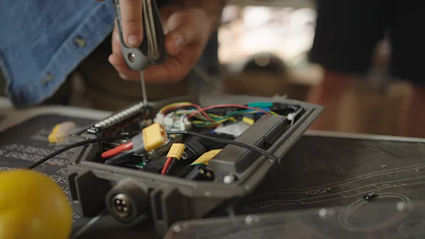Close-up of hands using a multi-tool on an opened Onewheel controller box, showing exposed wiring in red, yellow, green and blue and connectors on a workbench.