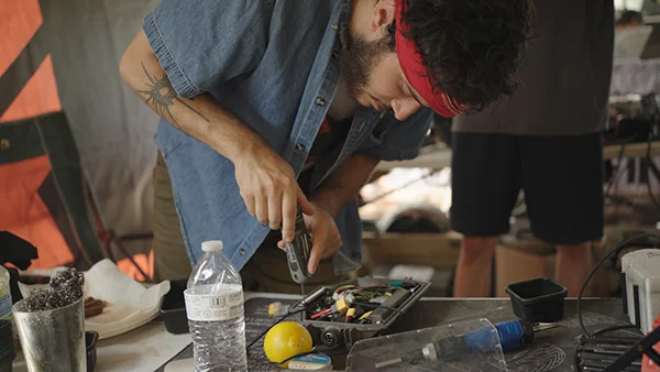 Jay leans over a workbench under a canopy tent at Oak City Shred Fest, working on electronic components laid out in a parts tray.