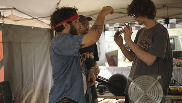 Jay reaches up to hand a component to another rider under the Fungineers tent while a third person stands nearby.