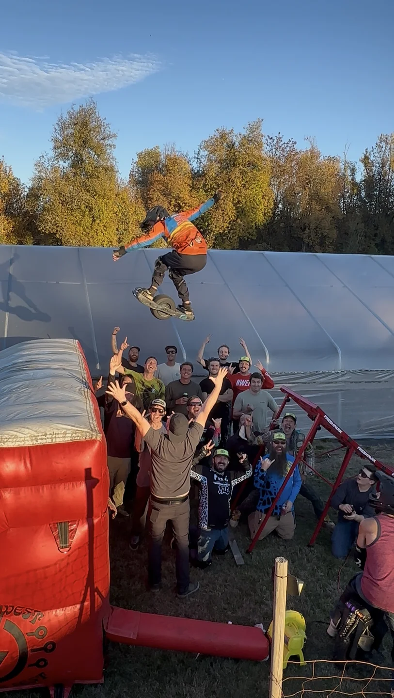 A Onewheel rider airborne well above head height over a crowd of spectators with their arms raised, coming down toward a red inflatable landing bag