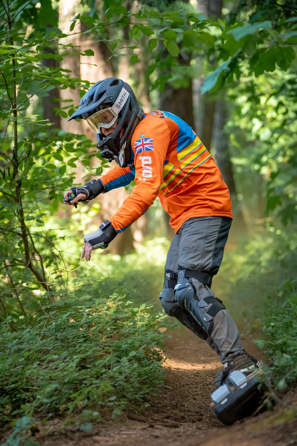 A Onewheel rider carving forest singletrack in a full face helmet with goggles and a chin-mounted camera, orange and blue jersey, wrist guards, moto pants and knee braces