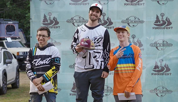 Three Onewheel racers in front of a Seek n' Shred ShredFest backdrop, the third-place rider on the right in an orange, blue and yellow Dave Ford Motorsports jersey making a peace sign