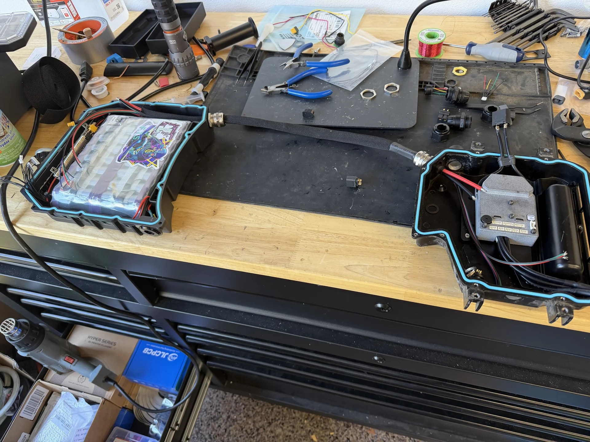 Two opened Onewheel enclosures on a workbench, one holding a battery pack and one holding a controller, joined by a sleeved cable, surrounded by hand tools and solder