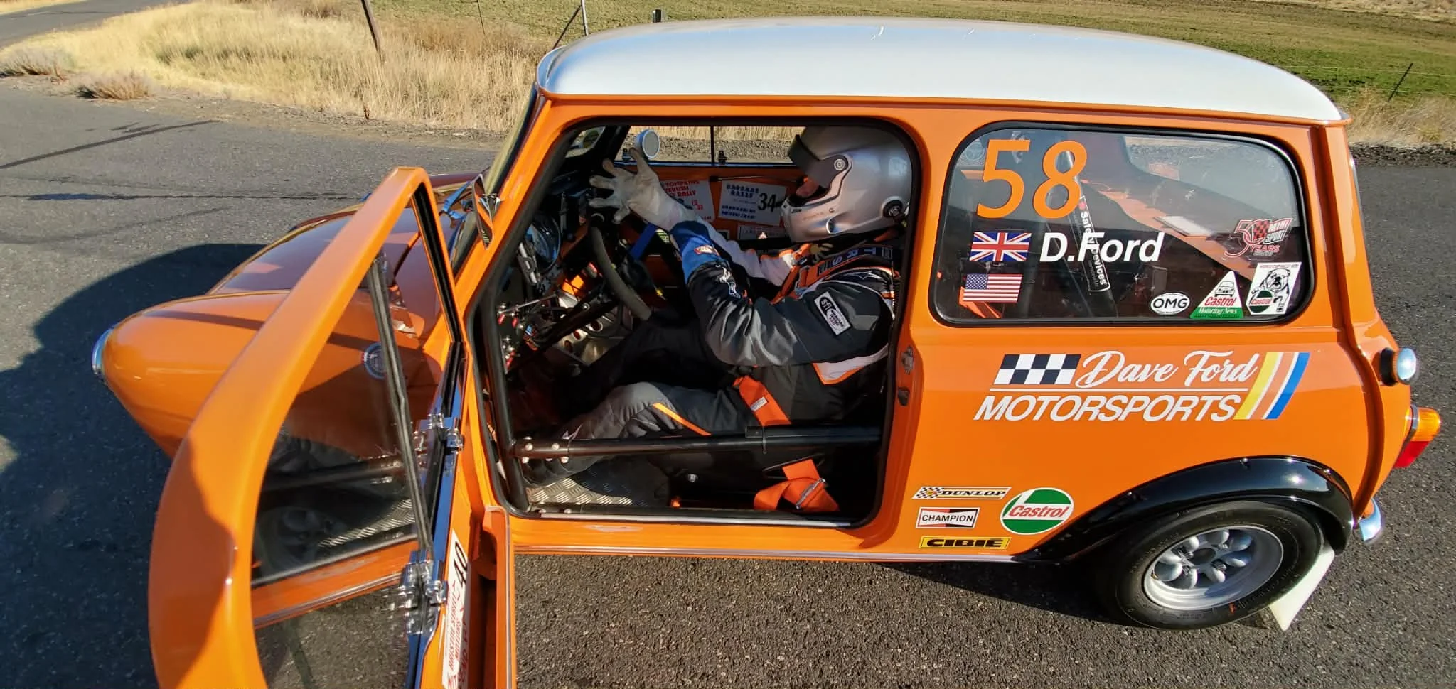 A man in a racing suit and flat cap standing beside an orange classic Mini with a white roof and Dave Ford Motorsports livery, at a paddock with dry hills behind