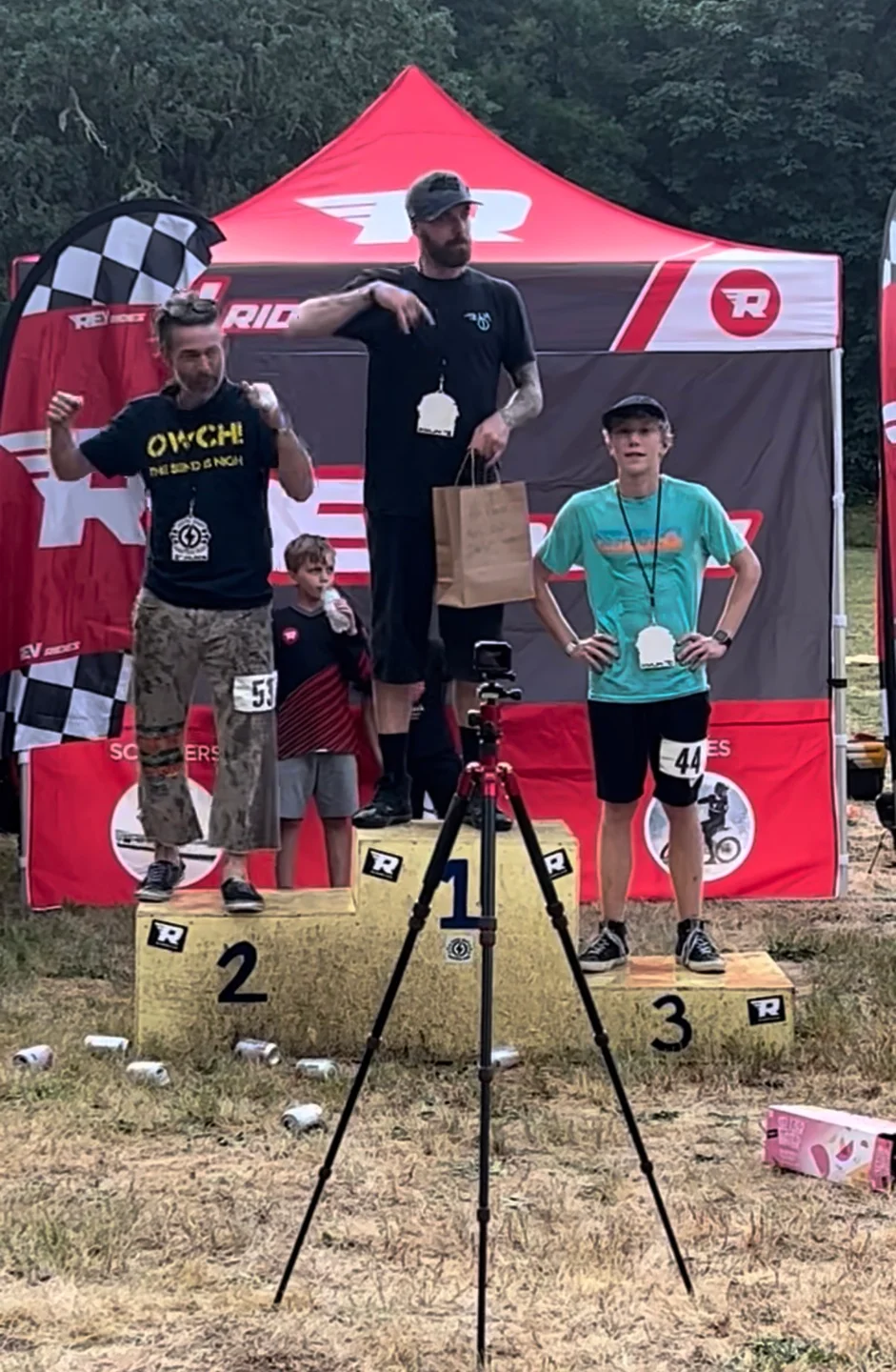 Three riders standing on numbered podium boxes under a red Rev Rides canopy at an outdoor race, the third-place rider wearing bib 44